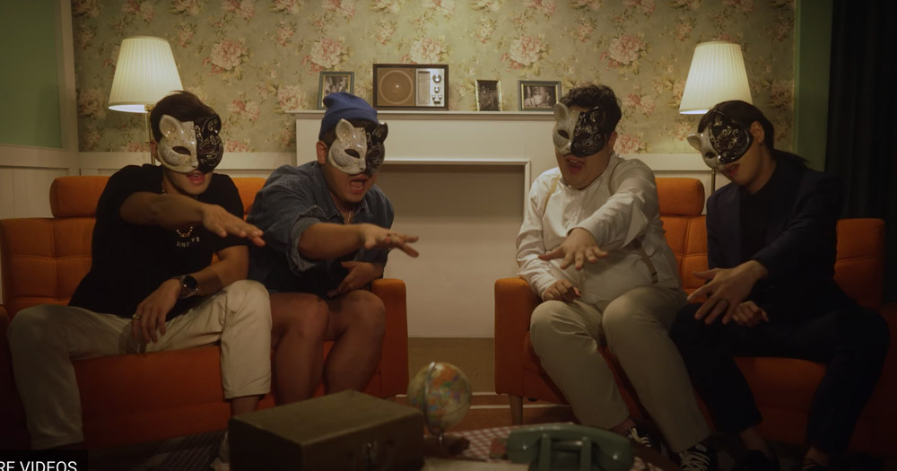 Screenshot from Lionesses, LGBTQ+ K-pop boy band. Sitting room scene with four men wearing cat masks waving hands in front.