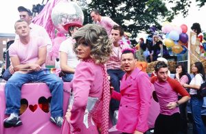 Photograph of a Pride parade with people and float dressed in pink. The central figure is the drag queen Panti Bliss. She is standing in profile looking over her shoulder towards the camera.