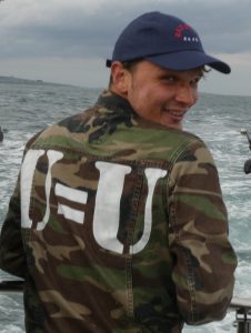 Mid length portrait of the back of a man, Robbie Lawlor. He is wearing a camouflage with the letters U=U in white. He has head head turned over his shoulder to look at the camera and is wearing a navy baseball cap.
