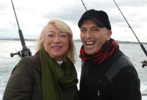 Mid length picure of woman on left, Anne Doyle, and Veda on right. In the background is the sea and fishing rods.