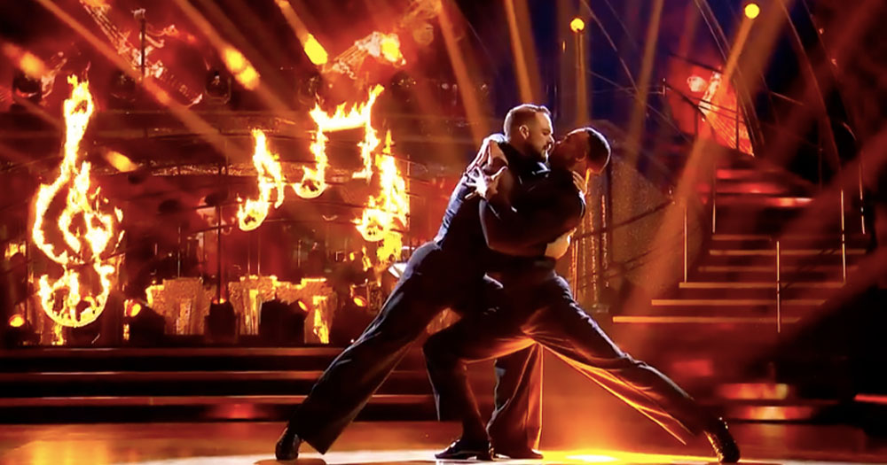 John Whaite and Johannes Radebe in a tango position on a stage with a background of flames in the shape of music notes. The pair is preforming on a Strictly Come Dancing episode.