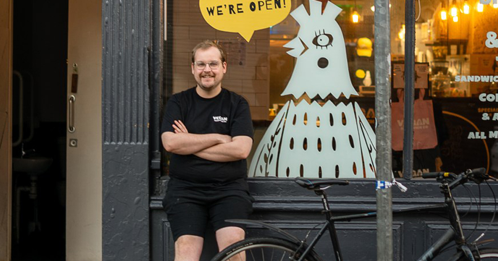Vegan Sandwich Co owner, Sam Pearson, sitting outside the shop on Queens Street in Dublin. He is resting against the window with his arms folded.