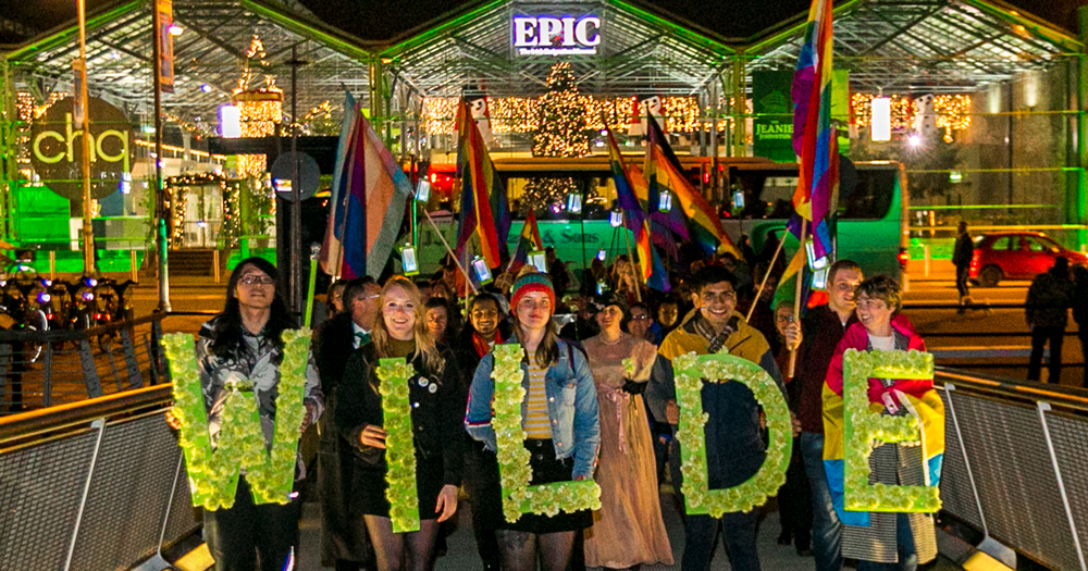 A group of people during Winter Pride holding Pride flags and a banner which spells WILDE. Behind them, the Epic museum