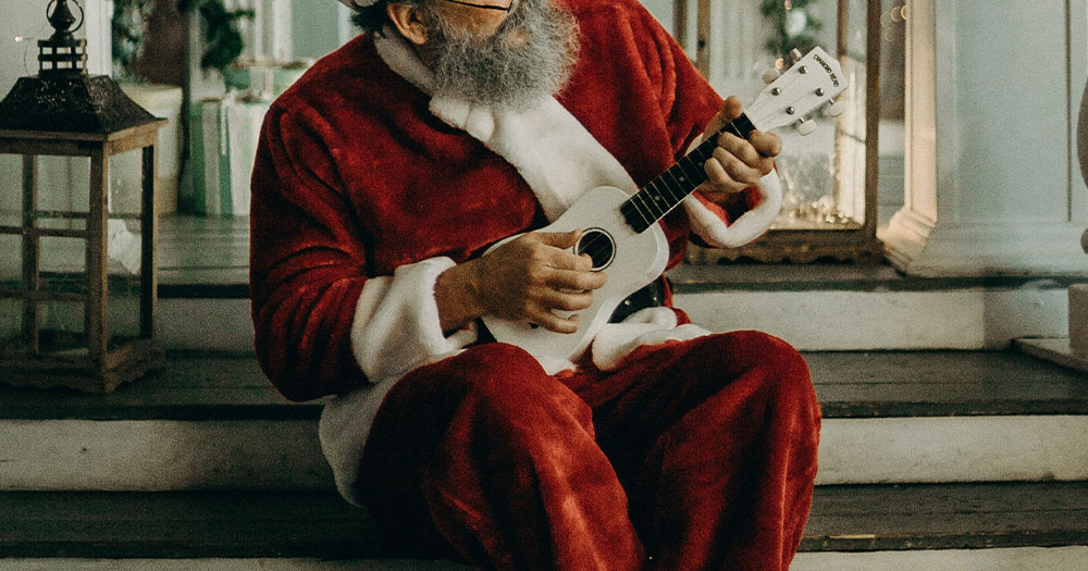 A man in a Santa Claus costume sitting on the door way playing a ukulele. 11 queer Christmas jams to add to your playlist
