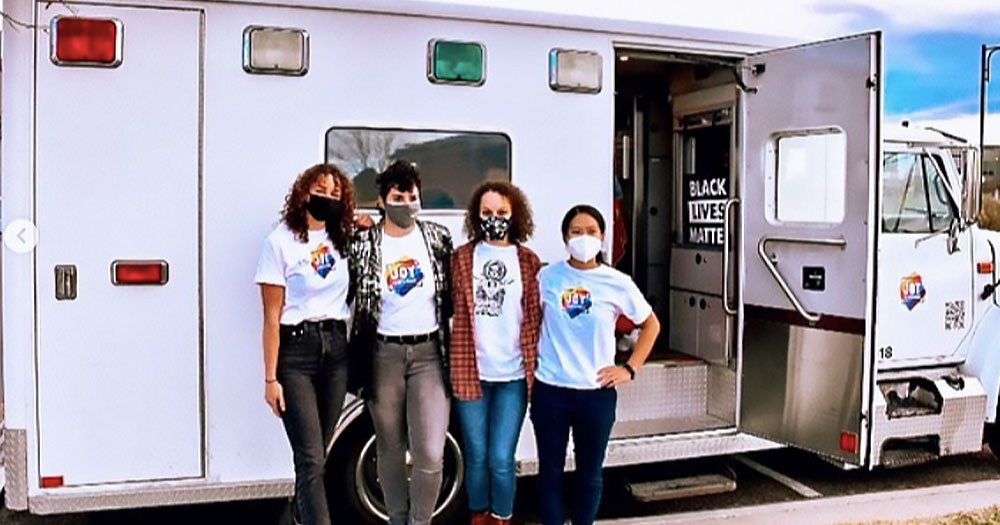 Four women in masks stand in front of a white ambulance. Retired ambulance transforms into a mobile mental health clinic for LGBTQ+ youth in Denver.