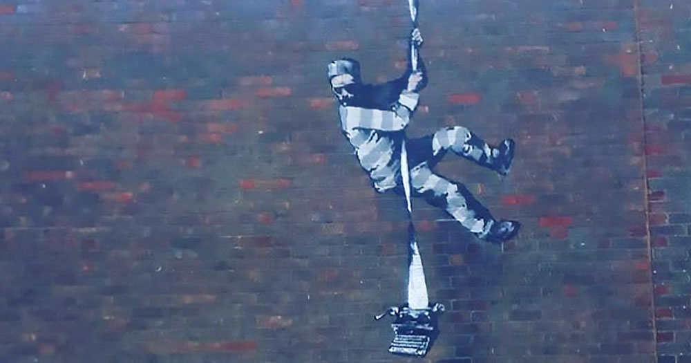 Painting on a brick wall of a prisoner climbing down a rope with a type writer. Banksy pledges £10m for the stencil of this street art to secure the purchase of Reading Gaol prison.