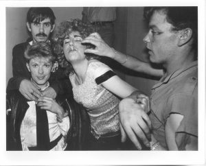 Black and white photograph of four clubbers from 1987. One man to the left is trying to remove a cigarette from a girls mouth in the centre of the image. The other two people are standing arm-in-arm to the left looking into the camera.