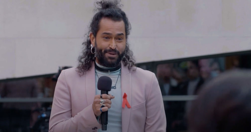 Film still of Luis Noguera Benitez from the film Positive Storytelling. Luis is holding a micraphone and is wearing a light pink jacket with a red AIDS ribbon on it. He has long hair tied in an upward ponytail.