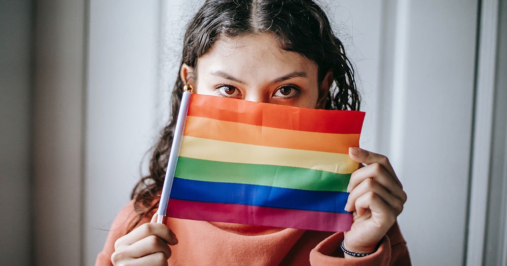 Girl's eyes looking directly at viewer while holding rainbow flag in front of the rest of her face. These are the latest guidelines on LGBTQ+ staff and students in Victoria and New Zealand.