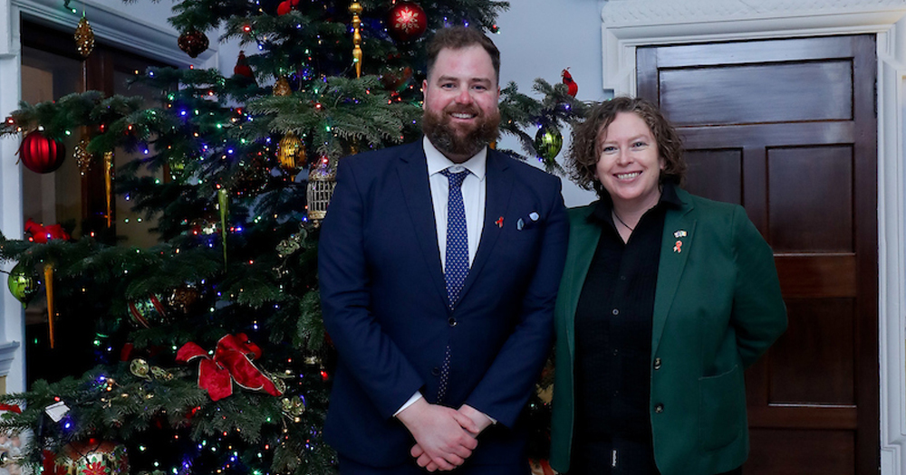 Loretta Cosgrove and Brian Murphy from Sydney Queer Irish Standing beside a Christmas tree. Brian is in the centre wearing a navy suit. Loretta is to his right wear a black shirt and emerald green blazer unbuttoned.