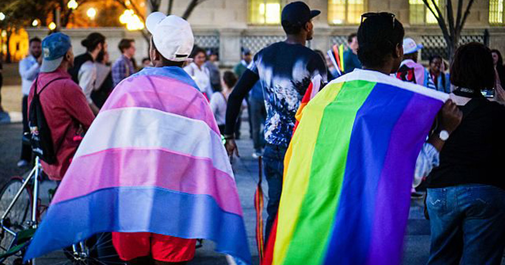 Together Conference - Photograph of two young people shot from behind. The one of the left has a Trans flag draped on their shoulders and the one on the right has a rainbow flag on their shoulders.