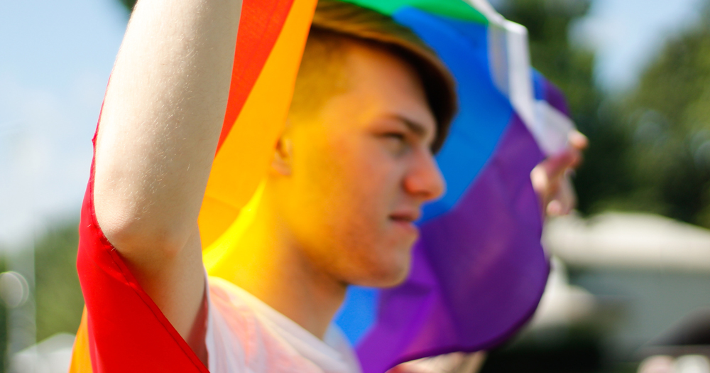 Young man, holding a LGBTQ+ Pride flag above his head in a display of queer joy