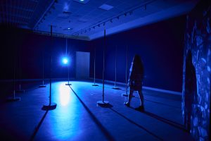 Photograph of the outside of the Array Collective installation. There is a circle of 9 flagpoles bathed in a blue light. A woman is walking through the circle.