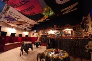Photograph of the inside of the Array Collective installation. It is a replication of an Irish síbín. The room has a bar on the right side of the room. There are three tables spread around the centre and along the left wall is a bench. The ceiling and walls are adorned with various banners, placards with left-wing political slogans.