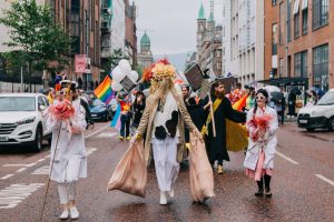 Array Collective at the Pride March 2019. In the photograph several people are dressed in outlandish costumes walking down a deserted street. The person in the middle is wearing a long beige trench coat open with white loose clothing underneath. Blonde hair covers their face and they have a headdress of yellow Dahlias. Their arms are outstreched and they have two heavy beige coloured bags in each hand.