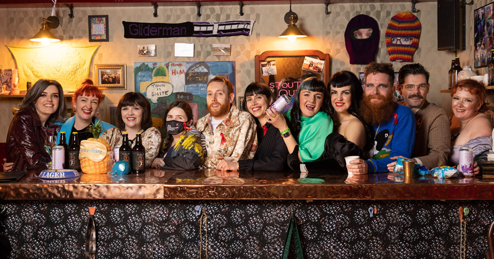 Photograph of the 11 members of Array Collective standing behind the bar of their Síbín installation as part of the Turner Prize exhibition.