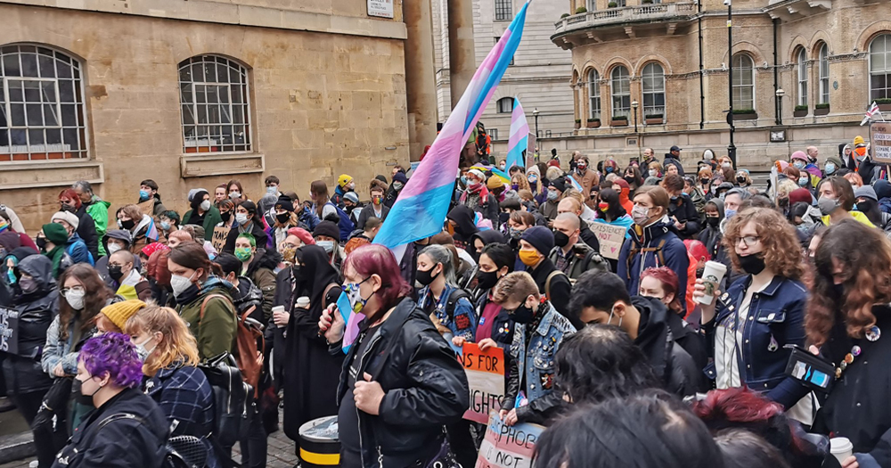 BBC Protest - Photograph of hundreds of people demonstrating outside BBC London head quarters with a furled Trans flag in the centre of the image.