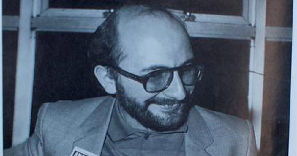 Black and white photograph of Charles Self. He is wearing an open blazer with a polo shirt underneath. He is looking downwards to the right and smiling. He is bald with a beard and glasses.