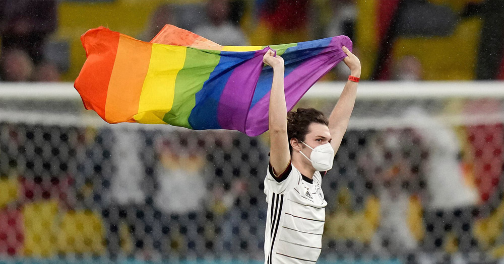 Photograph of a man wearing a white and black football jersey and a white face mask, running passed a soccer goal waving a Pride flag.