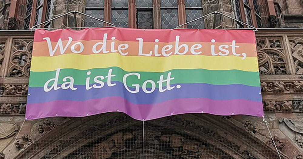 Photograph of a Pride banner hanging in front of a Church. The words 