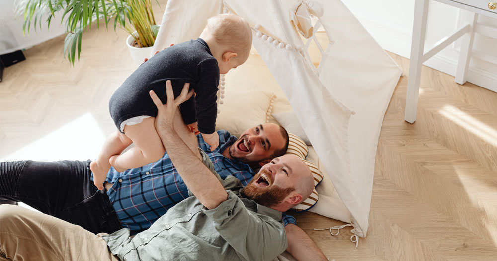 Growing Families: a pair of men are lying on the ground in a homemade fort holding up their young child