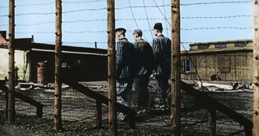 Three figures, one in uniform, one in concentration camp rags walk through Auschwitz with their backs to the camera