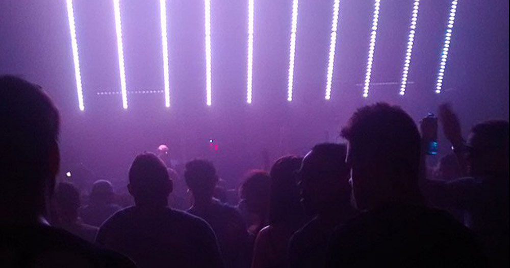 Photograph from a queer club. The scene has shadowed heads of people with lavender strip lights over head.