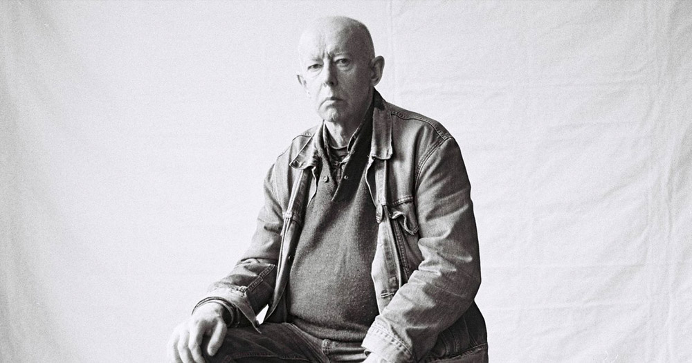 Black and white portrait photograph of Oliver Hegarty. He is sitting with his hands resting on his thighs wearing an open denim jacket with a sweatshirt underneath. He has a serious expression and is bald.