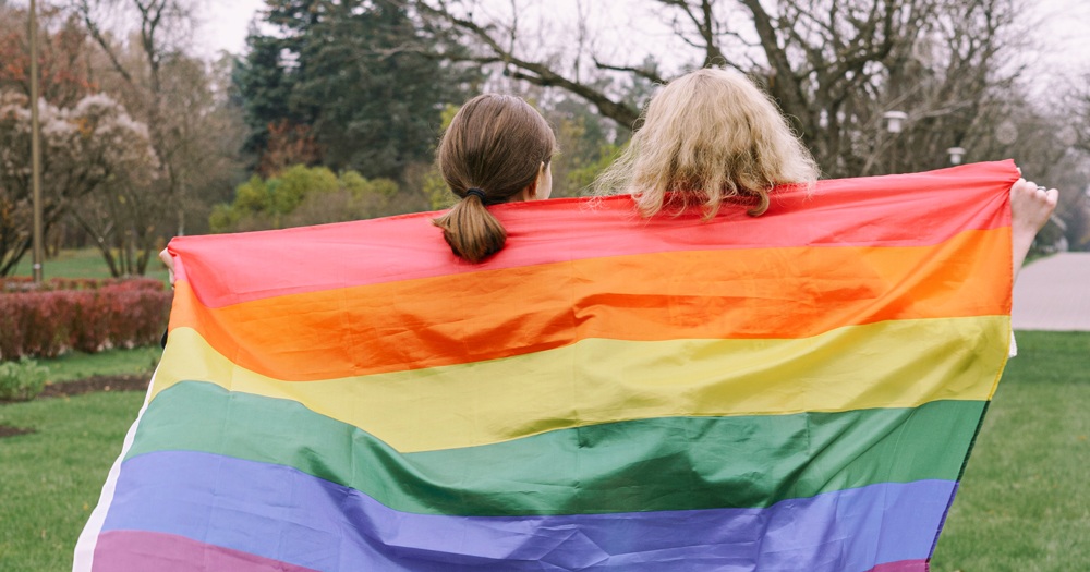 Two young women seen from behind wrapped in a rainbow flag