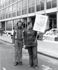 Black and white photo of a man and a woman standing side-by-side. The woman is holding a sign with reads, Lesbian Love.