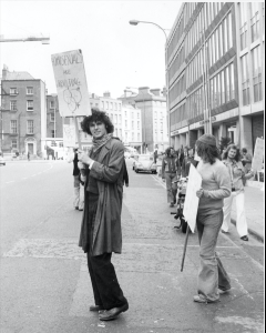 Black and white photo of a protest march. The man in front is facing the camera holding a placard which reads, Homosexuals are Revolting.