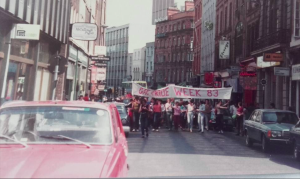 Colour photograph of of protesters marching in the background with a red car in the foreground.