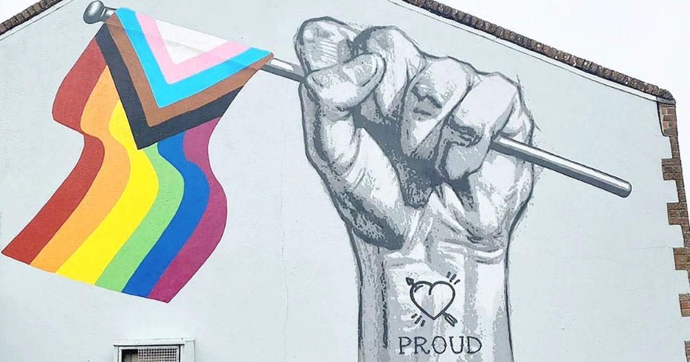 Photograph of new mural in Portlaoise. The painting shows a raised fist waving a progressive Pride flag. On the inside of the wrist is a heart and arrow tattoo with the word 'Proud' underneath.