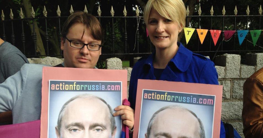 Photograph of Adam Long and Averil Power outside the Russian Embassy in Dublin. They are holding up posters of Putin with a rainbow coloured Hilter-style moustache.