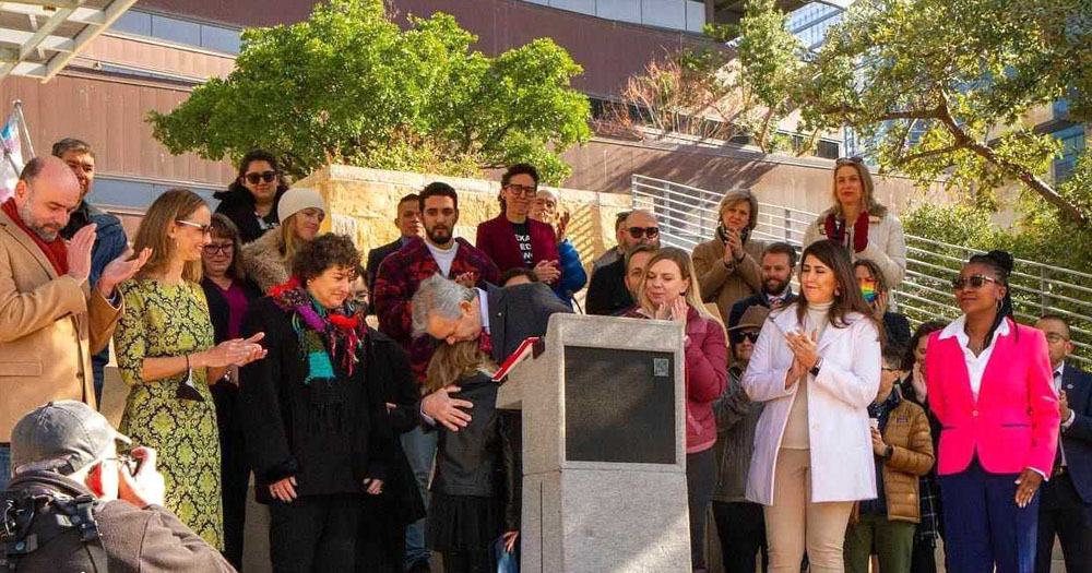 Photograph of Mayor of Austin, Texas, Steve Adler, hugging 11 year old Trans girl, Kai Shappley at the announcement of 'Transgender Youth and Family Safety Day'. They are standing at an outdoor podium surrounded by about 30 people.