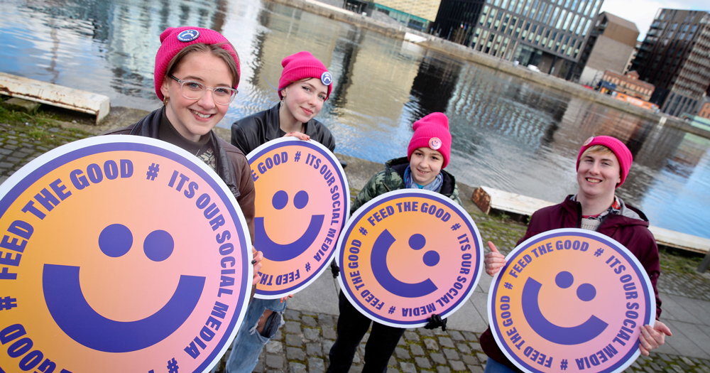 Four young people wearing pink hats holding round sings with smiley faces on. Young Irish LGBTQ+ people demand safer social media platforms through a new BeLonG To campaign