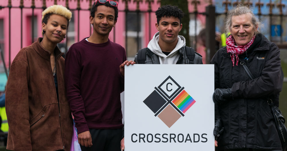 Iris Aghedo, Thomas Heising (Researcher), Leo O’Mahony, Ailsa Spindler (Gay Project Co-ordinator) at the launch of the Crossroads Report. The four people are photographed in a park with a pink and blue building in the background. They are holding a white card with the report logo.
