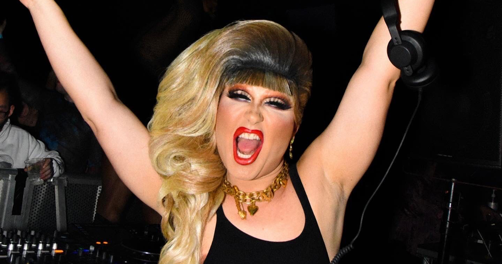 Drag queen Jodie Harsh wearing a blond wig and black dress cheering with her arms in the air while holding headphones