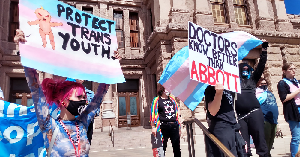 People protesting in Texas, where a judge blocked investigations into parents of Trans kids. They carry a Trans flag and signs reading 'Protect Trans Youth' and 'Doctors know better than Abbott'.