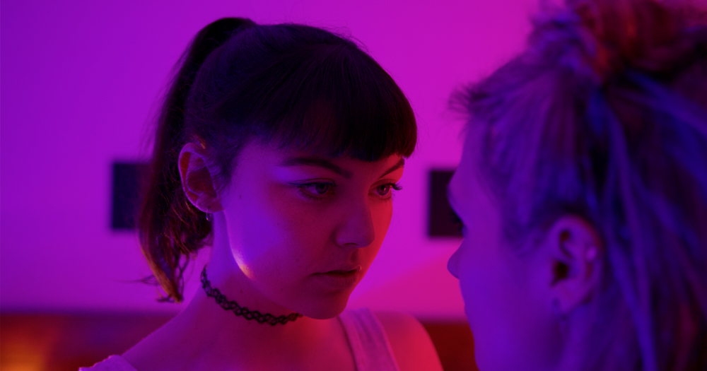 Still from the film Who We Love. The image shows the character of Lily in the main frame. She is looking intensely into the eyes of another woman who's facing away from camera to the right of the shot. The room is lit in pink and purple light.