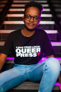 A model looking at the camera wearing a black T-shirt from the new GCN merch that reads 'Love your local queer press'.