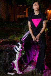 A model looking at the camera wearing a black T-shirt with a big pink triangle and has a black and white dog on a leash.