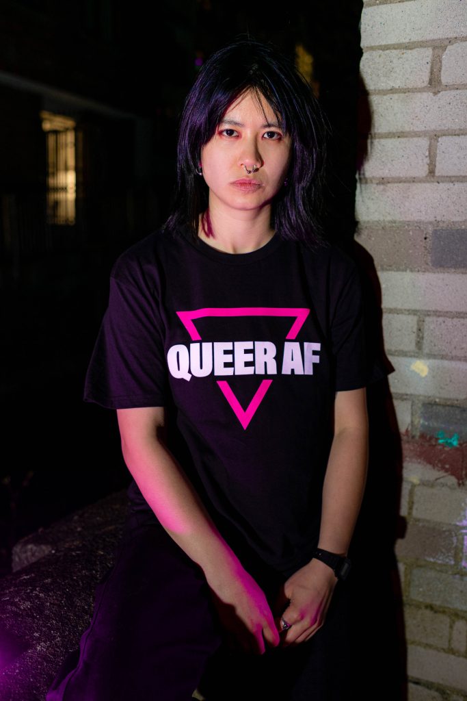 A model looking at the camera wearing a black T-shirt from the new GCN merch that reads 'Queer AF' with a pink triangle.