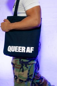 A model carrying a black tote bag that reads 'Queer AF'.