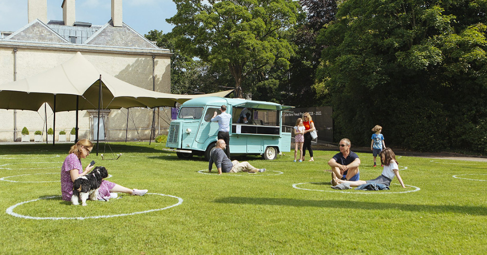 A picture of green grass and people sitting on it. It is a sunny day and there is a light blue coffee truck behind the folks on the grass.