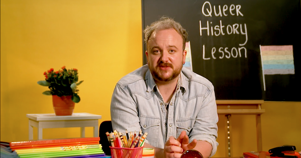 Shane Daniel Byrne sits at a desk with rainbow colored office supplies and a blackboard that says "Queer history lesson" behind him.