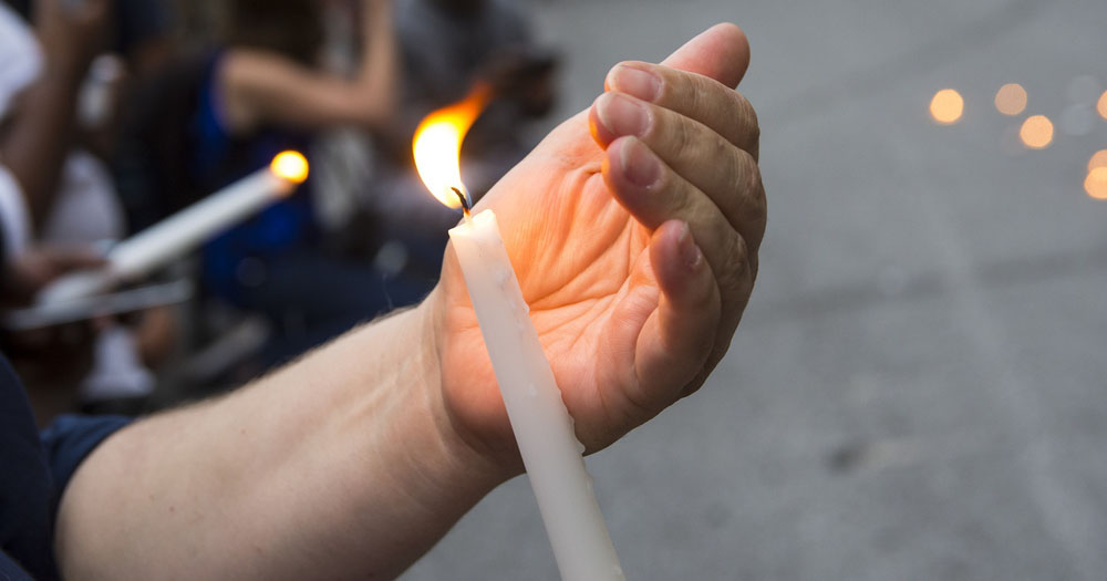 A photograph of a hand cupping the flame of a candle at a vigil.