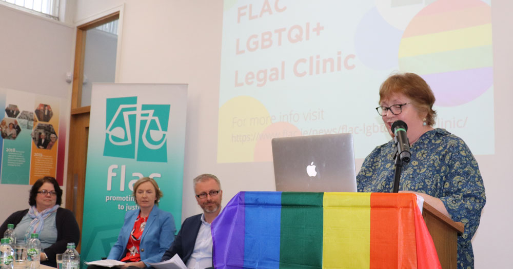 Photograph from the Launch of the FLAC LGBTQI+ Legal Clinic. Standing in front of a podium draped in a rainbow flag in FLAC CE, Eilis Barry. Seated to the right of her in left to right order are, Dr Mary McCauliffe, Paula Fagan and Minister Roderic O'Gorman.