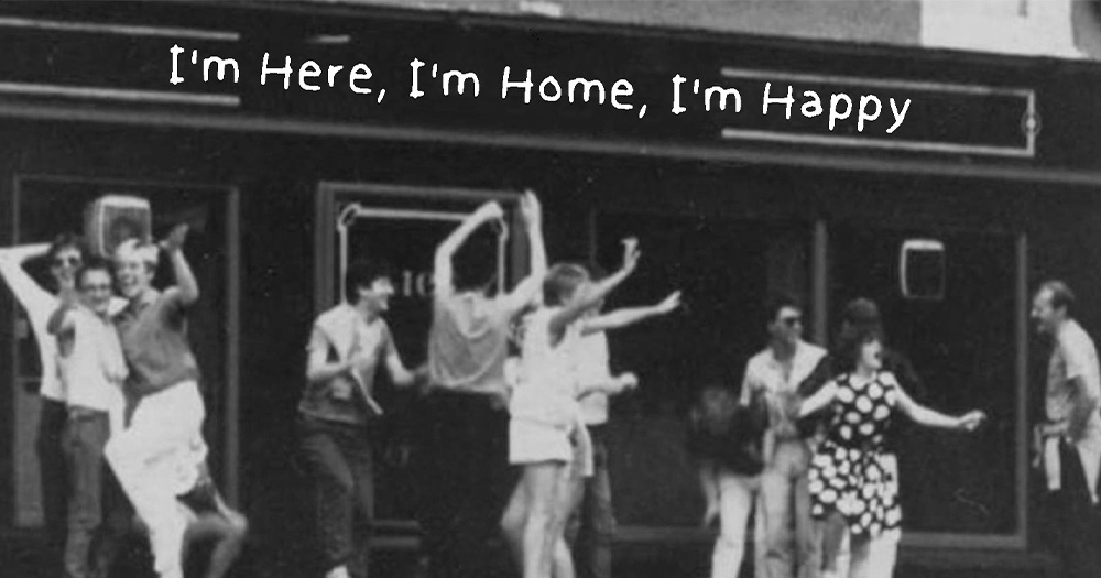 A black and white image of people dancing outside of a building. There is white text on the building which reads the name of the Cork LGBTQ Film I'm Here, I'm Home, I'm Happy.