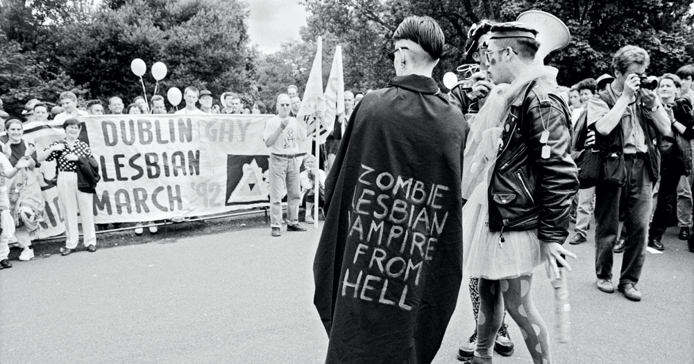 Black and white photograph of a protest march. In the foreground of the picture is a butch woman with her back to the camera. She is wearing a cape which reads 
